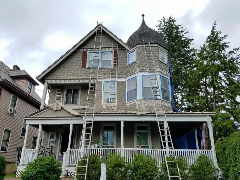 Gray shingle Victorian with a turret, three ladders against the facade and every window masked in blue tape before painting